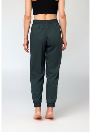 Naine seisab paljajalu selg vastu vaatajana, taljes elastsed tumesinised sportpüksid ja must varrukateta lühike top põhilisel taustal.