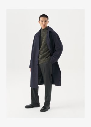 Marineblauer Mantel mit Knopfverschluss, grünem strukturiertem Pullover, schwarzen Hosen und schwarzen Lederstiefeln. Glatte Stoffqualität mit klarer Silhouette.