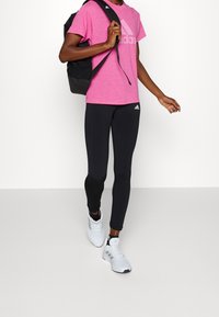 Camiseta rosa de Adidas con logo, leggings negros y zapatos deportivos blancos. Persona lleva una mochila negra, caminando frente a un fondo blanco liso.