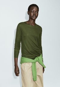 Maglia a maniche lunghe verde oliva in morbido materiale, abbinata a un maglione verde chiaro legato in vita e pantaloni beige a gamba larga.