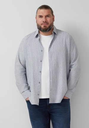 Bärtiger Mann mit langen nach hinten gebundenen Haaren trägt ein hellgraues Hemd über einem weißen T-Shirt und blauen Jeans, steht mit den Händen in den Taschen.