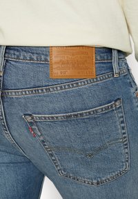 Gros plan de la poche arrière d’un jean Levi’s bleu avec un patch en cuir marron de la marque et une étiquette logo en tissu rouge, porté avec une chemise beige.