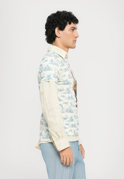 Man in een beige lange mouwen shirt onder een korte mouwen met patroon, met lichtblauwe streepjesbroek, staande in profiel tegen een witte achtergrond.