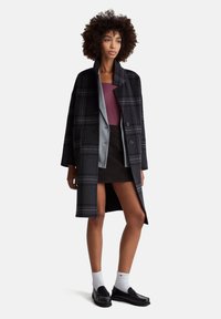 Frau trägt einen langen dunklen karierten Mantel über einem grauen Blazer, einem lila Oberteil, einem schwarzen Rock, weißen Socken und schwarzen Loafers, steht vor einem weißen Hintergrund.
