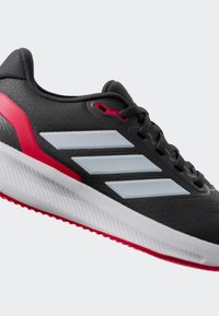 Chaussure de sport noire avec tige en mesh, présentant trois rayures gris clair, des accents rouges et une semelle blanche texturée avec un design cranté.