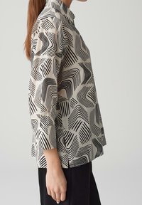 Blouse à motifs beige avec des dessins de vagues noires ; elle présente un col et des manches trois-quarts, fabriquée en tissu léger.