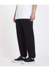 Pantalons de survêtement noirs avec une coupe fluide, fabriqués en tissu doux, avec un petit logo blanc sur le côté. Associés à des baskets noires.