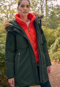 Parka verte avec capuche bordée de fourrure, portée par-dessus un gilet matelassé orange vif. Comprend des poches zippées et une coupe longue et droite. Cadre extérieur.