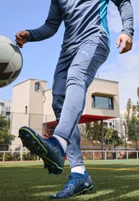 Tuta atletica blu con dettagli testurizzati, abbinata a calzature blu scuro con accenti verdi e borchie, posizionata su un campo erboso.