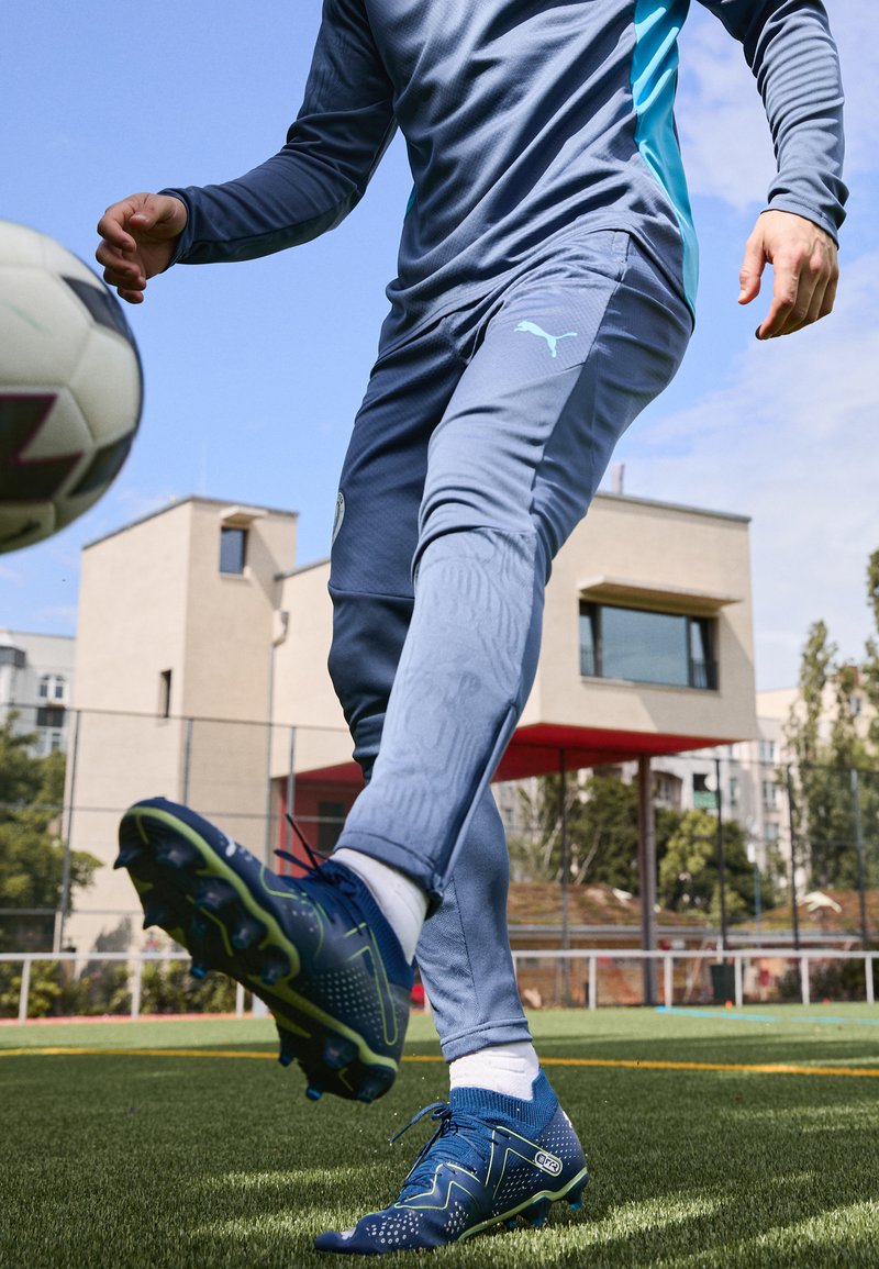 Tuta atletica blu con dettagli testurizzati, abbinata a calzature blu scuro con accenti verdi e borchie, posizionata su un campo erboso.