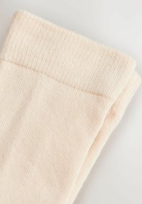 Krém színű bordázott kötött anyag, amelyet hajtva mutatják be, kiemelve a mandzsetta részletét egy puha, világos háttéren.
