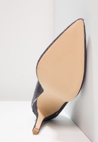 Svart strukturerad högklackad sko med spetsig tå. Platt beige sula med horisontella spår. Positionerad mot en vit bakgrund.