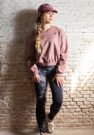 Roze sweatshirt met geribbelde boorden, gecombineerd met zwarte tie-dye leggings. Witte sneakers en een bordeauxrode pet maken de outfit af. Textuurmuur als achtergrond.