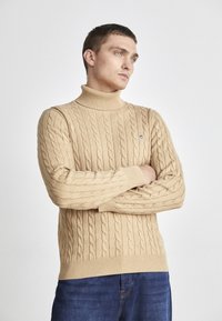 Jersey de cuello alto de punto beige con pu�ños y dobladillo acanalados, que presenta un pequeño logo en el pecho. Combinado con jeans azules.