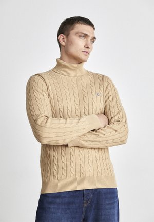 Beige kabelstrikket turtleneck genser med ribbestrikkede ermer og nederkant, med en liten logo på brystet. Brukt sammen med blå jeans.