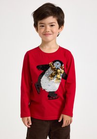 Red long-sleeve shirt featuring a sequined penguin design, wearing a yellow scarf and black skates. The penguin is multicolored and textured.
