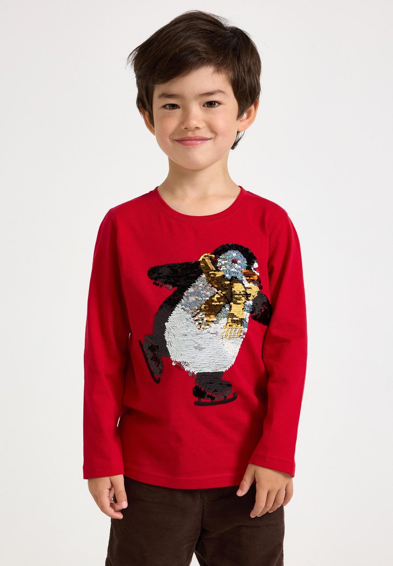 Red long-sleeve shirt featuring a sequined penguin design, wearing a yellow scarf and black skates. The penguin is multicolored and textured.