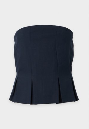 Mujer con el pelo castaño liso que lleva un top peplum negro sin tirantes y pantalones anchos de pierna en color oliva, de pie frente a un fondo blanco liso.
