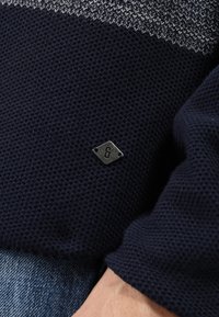 Mörk marinblå strukturerad stickad tröja ärm med en liten metallisk diamantformad logotagg ovanför blå denimjeans.