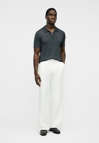 Muž oblečený v tmavě zeleném krátkém polosetru se zipem, bílých širokých kalhotách a černých botách, stojící s jednou rukou v kapse.