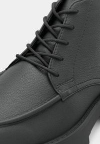 Gros plan d'une botte en cuir noir avec un bout arrondi, une surface texturée et des lacets ronds noirs passés dans des œillets métalliques.