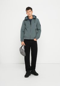 Graue Zip-Jacke mit Kapuze, schwarze Hose und schwarze Schuhe. Das Modell hält eine graue Umhängetasche. Hat einen gerippten Kragen und elastische Bündchen.
