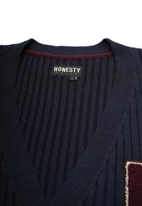 Marinblå ribbad cardigan med V-ringning, som har ett vinrött accentband vid kragen och en mönstrad patch på vänster sida. Storlek L-skylt synlig.