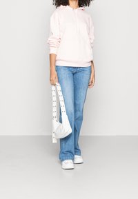 Donna in felpa rosa chiaro, jeans blu e sneakers bianche, che tiene una borsa bianca con tracolla borchiata su uno sfondo semplice.