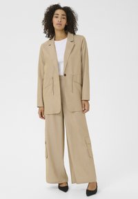 Beige Blazer mit Klappentaschen, kombiniert mit weiten Hosen mit seitlichen Cargo-Taschen. Beide Teile bestehen aus leichtem Stoff.