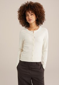 Krema krāsas kardigāns ar apaļu kakla izgriezumu, garām piedurknēm un pogu aizdari. Iezīmēts ar ribotu apakšmalu un piedurkņu galiem. Modelis nēsā tumšas bikses.