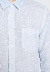 Jasnoniebieska, zapinana koszula z fakturowanej tkaniny, z białymi wzorami paisley i kwiatowymi, posiadająca kieszeń z przodu oraz kołnierz w szpic.