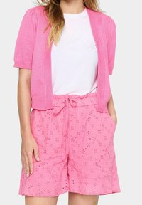 Vaaleanpunainen lyhythihainen neuletakki valkoisen t-paidan päällä, yhdistettynä vaaleanpunaisiin sortseihin, joissa on kirjontakuvioita ja joustavyötärö.