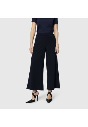 Frau trägt schwarze, weit ausgeschnittene, verkürzte Hosen, ein schwarzes Kurzarmoberteil und schwarze spitz zulaufende High Heels mit Knöchelriemen.