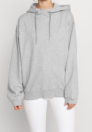 Personne portant un sweat à capuche gris clair surdimensionné avec cordons et un pantalon blanc, debout devant un fond clair uni.