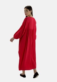 Rød, løstsiddende maxi kjole med struktureret stof, lange puffærmer og en samlet rygsøm. Båret med sorte slip-on sko.