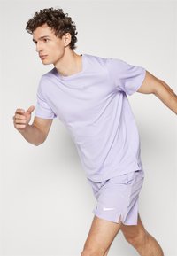 Šviesiai violetinė sportinė marškinėliai ir šortai, pagaminti iš lygaus, kvėpuojančio audinio. Marškinėlių ir šortų krašte yra subtilus Nike logotipas.