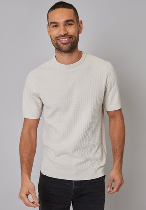 Suéter de punto de manga corta en color crema con un patrón texturizado, escote redondo y puños acanalados, combinado con jeans oscuros.