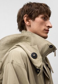 Beige trenchcoat med hög krage, texturerat material, svarta knappar och dragkedjestängning. Designen har synliga sömmar.