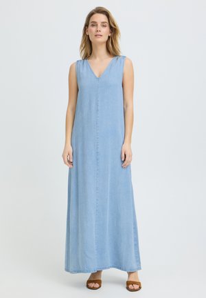 Naine seisab, kandes varrukateta helesinist V-kaelusega maksikleit ja pruune avatud varbaga sandaale ühtlasel heledal taustal.