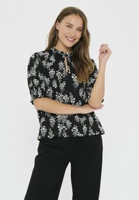 Blusa nera con motivo floreale bianco, maniche a sbuffo, scollatura a chiave e una vestibilità rilassata. Abbinata a pantaloni scuri, texture liscia.