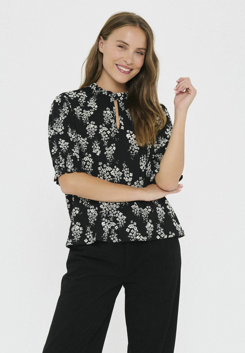 Blusa nera con motivo floreale bianco, maniche a sbuffo, scollatura a chiave e una vestibilità rilassata. Abbinata a pantaloni scuri, texture liscia.