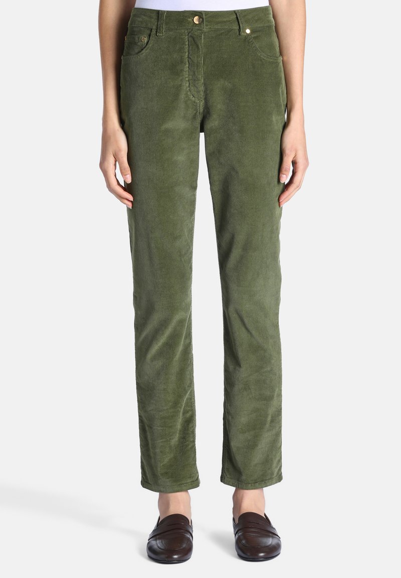 Femme debout portant un pantalon en velours côtelé vert à jambes droites et des mocassins brun foncé, mains reposant le long du corps sur fond blanc.