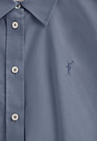 Camicia azzurro chiaro con texture intrecciata, caratterizzata da bottoni bianchi e una piccola figura ricamata che tiene una mazza da golf.