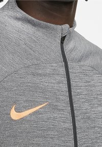 Gray zip-up jacket in textured fabric with a high collar, featuring a Nike logo in orange. Zipper closure along the front.