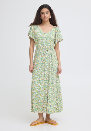 Femme aux longs cheveux bouclés portant une robe midi florale pastel et des tongs jaunes, debout devant un fond clair uni.