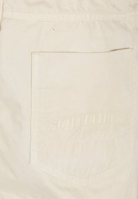Beige bomuldsstof med en påsyet lomme, der har prægede "SOUTHPOLE ORIGINAL" bogstaver. Glat tekstur, enkelt design og klassisk form.