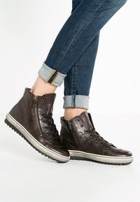 Hochtop-Sneaker aus dunkelbraunem Leder mit strukturierter Oberfläche, ausgestattet mit schnürbarem Design und seitlichen Reißverschlüssen, auf einer weißen Gummisohle.