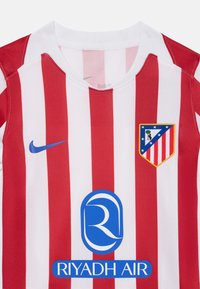 Crvena i bijela prugasta nogometna majica s okruglim izrezom, koja sadrži plavi Nike logo, plavi sponsor logo Riyadh Air i grb momčadi u obliku štita.