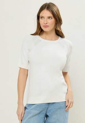 Kvinde iført en hvid kortærmet sweater og lyseblå jeans, stående mod en ensfarvet lys baggrund, kiggede til siden.