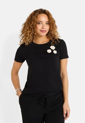 Camiseta negra de manga corta hecha de algodón, con tres aplicaciones florales en color crema en el lado izquierdo. Cuello redondo simple.